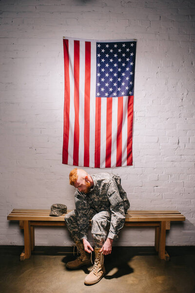 american soldier in military uniform tying shoelaces with flag on white brick wall behind, 4th july holiday concept