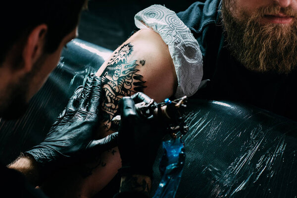 обрезанный снимок татуировщика в перчатках, работающего на татуировке на плече в салоне
