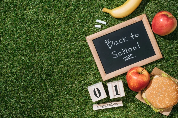 flat lay with blackboard with back to school lettering, calendar, banana, apples and burger on green grass