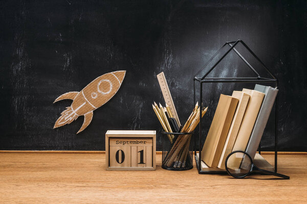 close up view of cardboard rocket on blackboard, calendar, magnifying glass and books on wooden tabletop