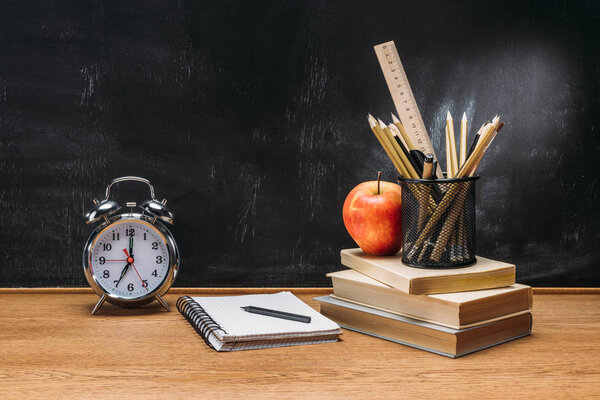 close up view of fresh apple, clock, notebook, pencils and books on wooden tabletop with empty blackboard behind