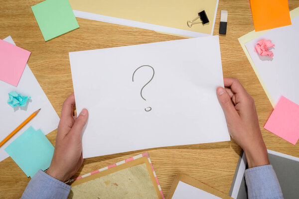 cropped image of woman holding paper with question mark over table with stick it notes and stationery supplies 