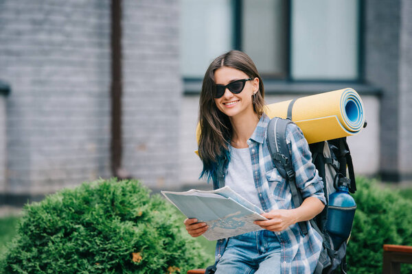 happy young woman with backpack holding map and smiling at camera