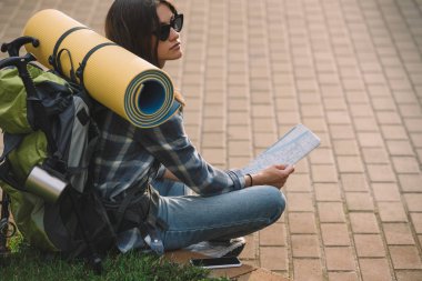 young female backpacker in sunglasses sitting on grass and holding map