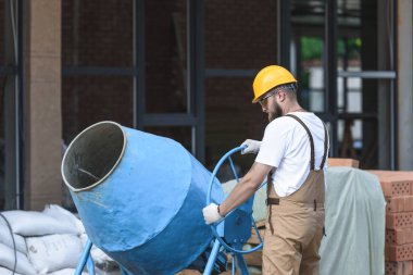 construction worker in hardhat and protective googles working with concrete mixer on construction site  clipart