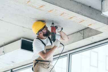 low angle view of builder in protective googles and hardhat working with drill at construction site  clipart