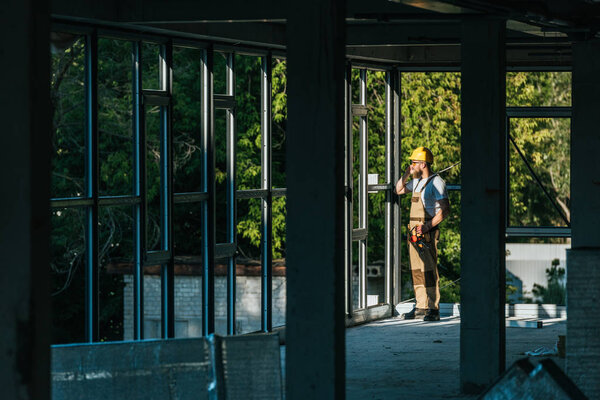 distant view of builder in protective helmet and googles talking on smartphone at concstruction site 