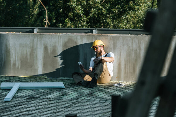 young builder in hardhat and protective googles using smartphone and resting at construction site 
