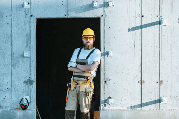  builder in protective googles and hardhat standing with crossed arms at construction site 