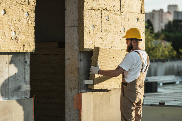 side view of builder in protective helmet and googles doing insulation of wall at construction site 