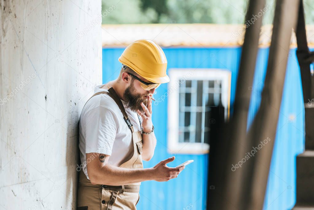 side view of builder in hardhat and protective googles smoking cigarette and talking on smartphone at construction site 