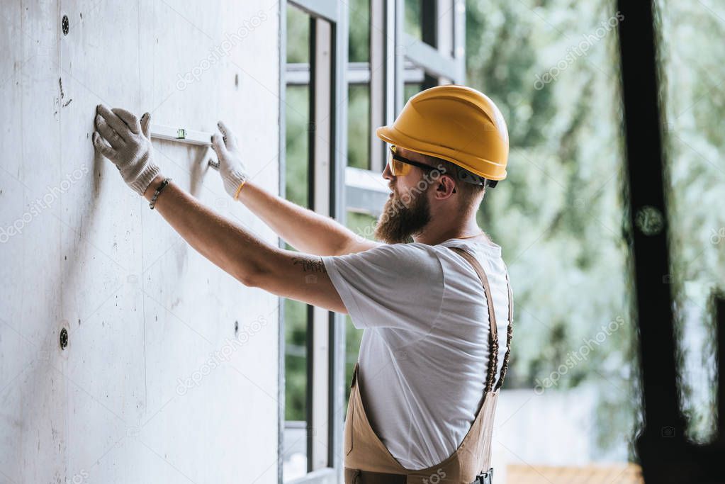 side view of builder in protective helmet and googles using spirit level at construction site 