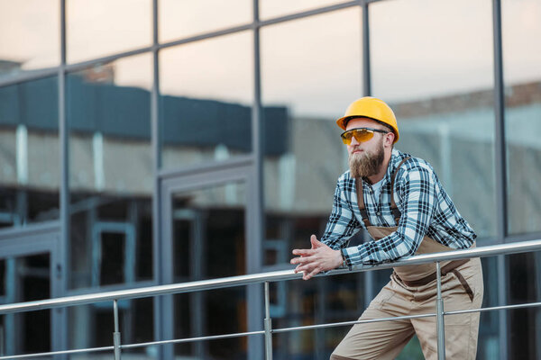 young builder in protective googles and hardhat holding hands together near building