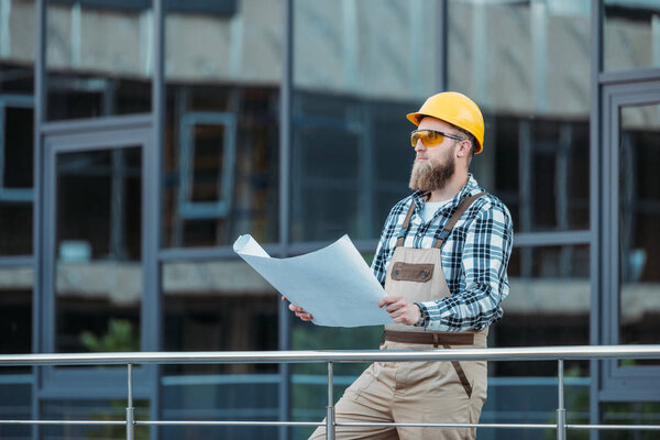 young construction worker in protective googles and hardhat looking at blueprint  