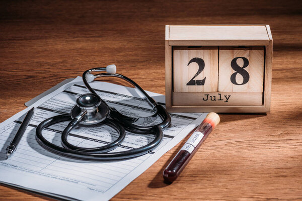 selective focus of test flask with blood sample, stethoscope, medical questionary and wooden calendar with 28th july date on table, world hepatitis day concept