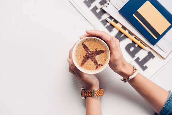 cropped shot of woman holding cup of coffee with plane sign at tabletop with credit card, passport and ticket, traveling concept