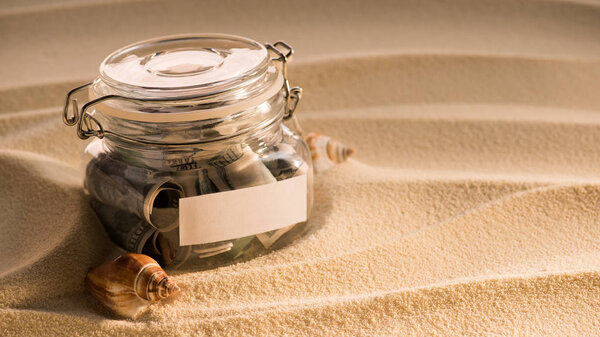 close up view of glass jar with dollars on sandy beach with seashells, traveling concept