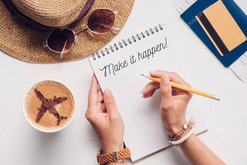 partial view of woman with notebook with make it happen lettering at tabletop with cup of coffee, straw hat, passport and ticket, vacation concept