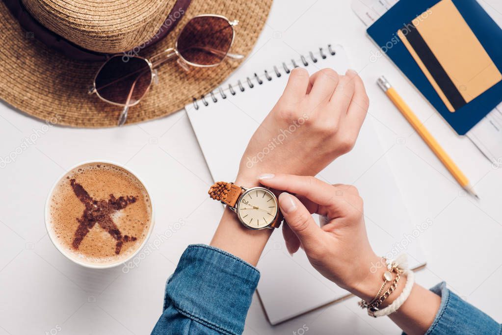 cropped shot of woman checking time at tabletop with straw hat, sunglasses, passport and notebook, traveling concept