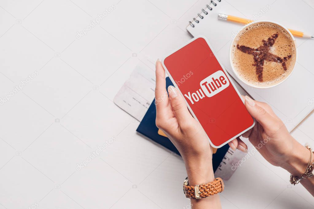 Cropped shot of woman with smartphone with youtube logo on screen at tabletop with cup of coffee, ticket and passport