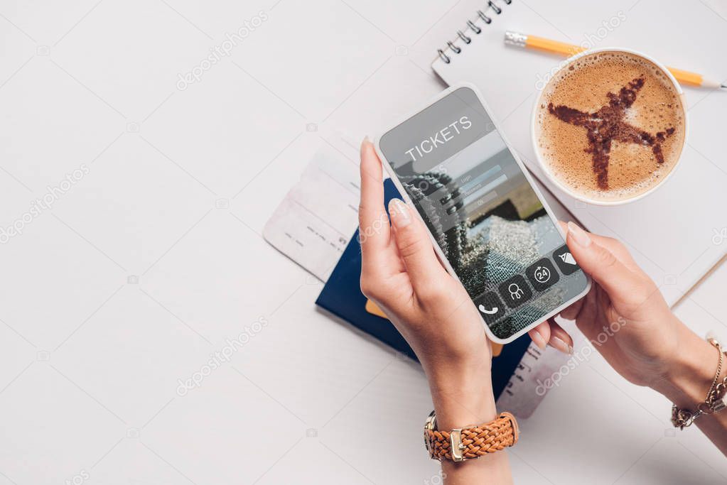 Cropped shot of woman with smartphone with tickets website on screen at tabletop with cup of coffee, ticket and passport