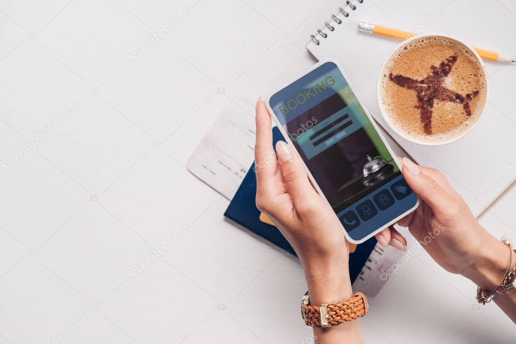 Cropped shot of woman with smartphone with booking website on screen at tabletop with cup of coffee, ticket and passport