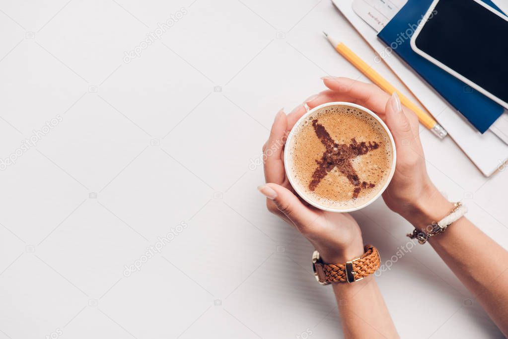 cropped shot of woman holding cup of coffee with plane sign at tabletop with passport, smartphone and ticket, traveling concept