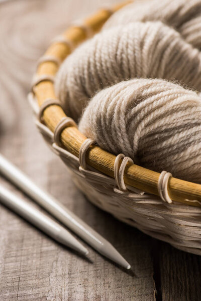 selective focus of beige knitted woolen yarn in wicker basket near knitted needles on wooden background 