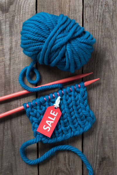 top view of blue knitting needles, blue yarn ball and sale tag on wooden background