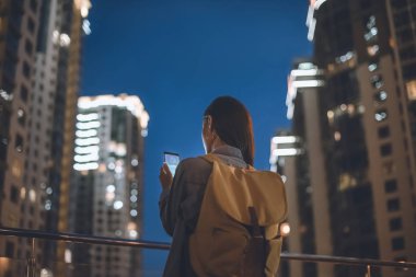 Arkadan Görünüş sırt çantası ile kadın ve logo üstünde perde gece şehir sokak üzerinde duran elinde alışveriş ile smartphone