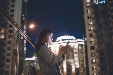 çekici kadın arka plan üzerinde gece şehir ışıkları ile sokakta smartphone kullanarak yan görünüm