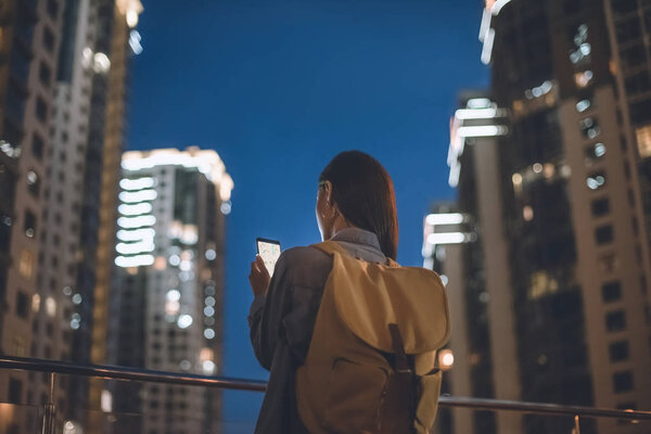 back view of woman with backpack and smartphone with map on screen in hands standing on night city street