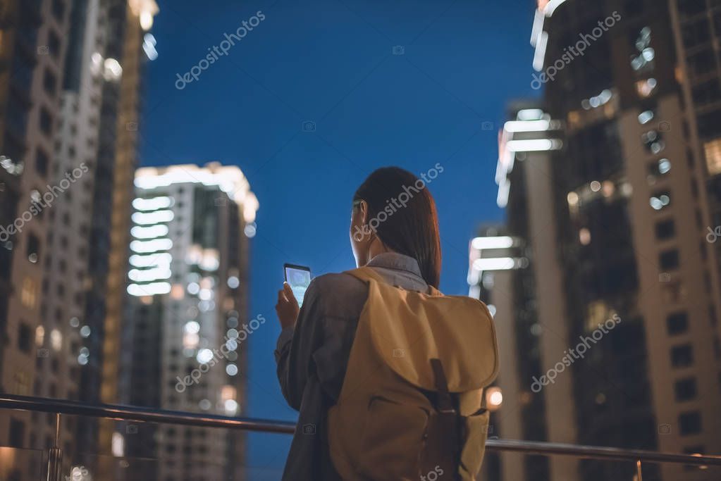 Back view of woman with backpack and smartphone with shopping logo on screen in hands standing on night city street