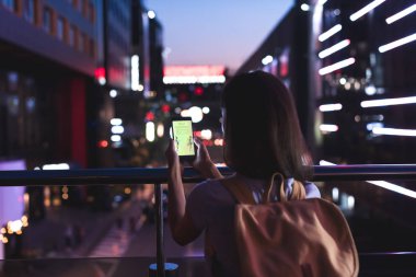 Arkadan Görünüş sırt çantası ile kadın ve Web sitesinde ekran gece şehir sokak üzerinde duran elinde alışveriş ile smartphone