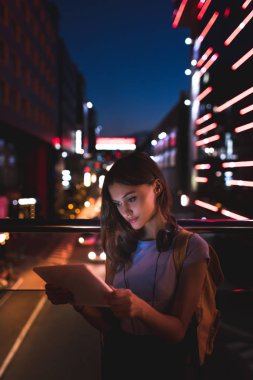 portrait of young woman with headphones using tablet on street with night city lights on background