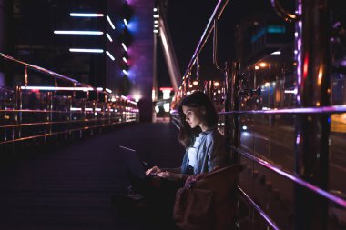 side view of young woman using laptop on street with night city lights on background clipart