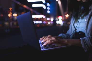 cropped shot of woman with headphones typing on laptop on city street at night clipart