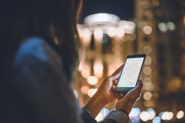 partial view of woman using smartphone with map on screen and night city lights on background