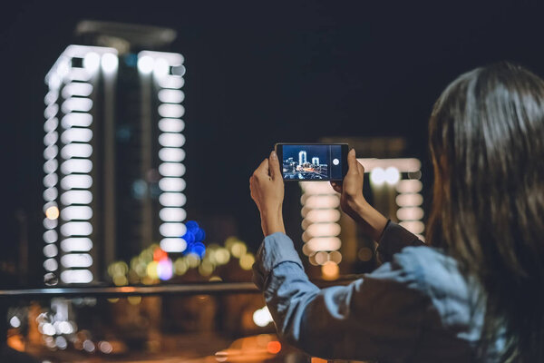 вид сзади женщины, фотографирующей город на смартфон в руках ночью
