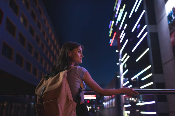 beautiful pensive woman with backpack looking away on city street at night