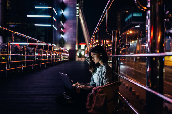 side view of young woman using laptop on street with night city lights on background