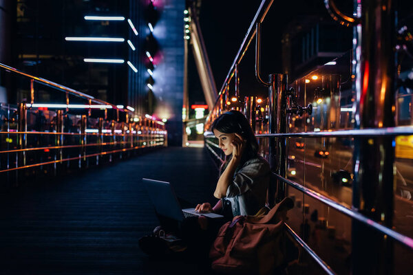 side view of woman listening music in headphones while using laptop on city street at night