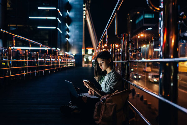 вид сбоку на молодую женщину с ноутбуком на коленях с помощью смартфона на фоне ночного города
