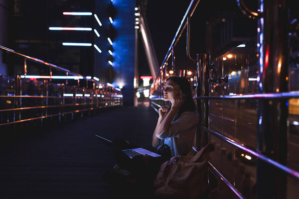 side view of woman listening music in headphones with laptop on knees on city street at night