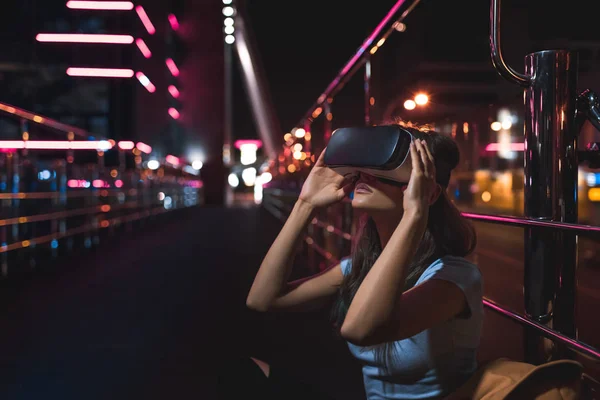 young woman in virtual reality headset sitting on street with night city on background