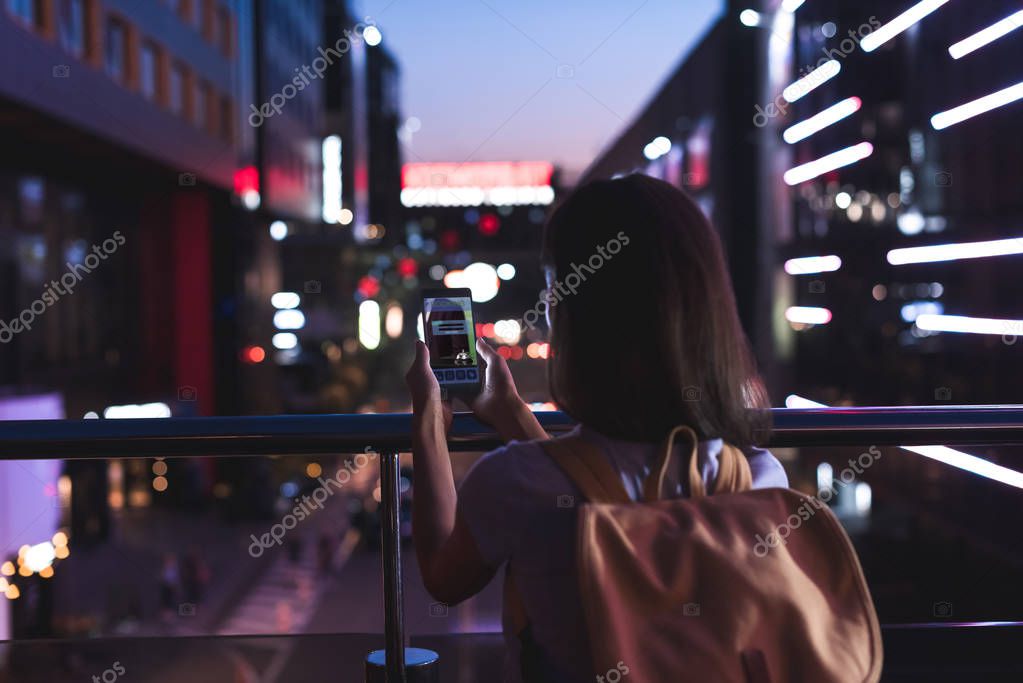 Rear view of woman with backpack and smartphone with booking website on screen in hands standing on night city street