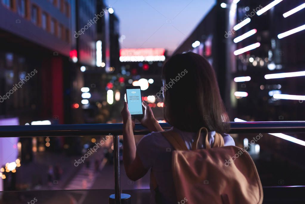 Back view of woman with backpack and smartphone with skype logo on screen in hands standing on night city street