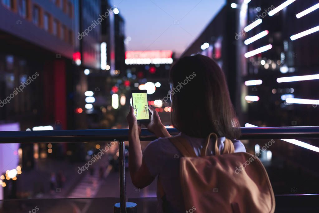 Back view of woman with backpack and smartphone with shopping website on screen in hands standing on night city street
