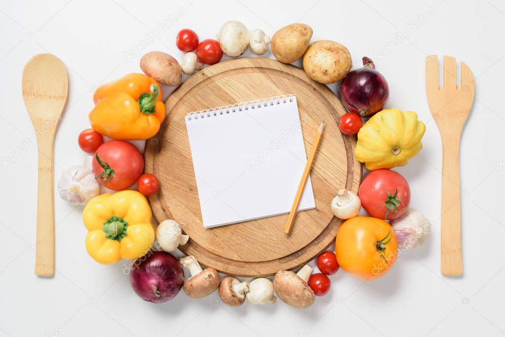 top view of ripe vegetables around wooden board, notebook and pencil isolated on white