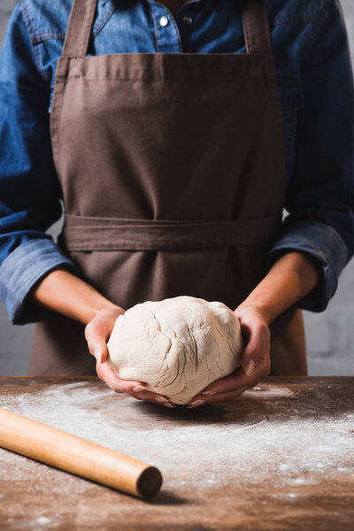 cropped shot of woman in apron holding raw dough for pizza in hands